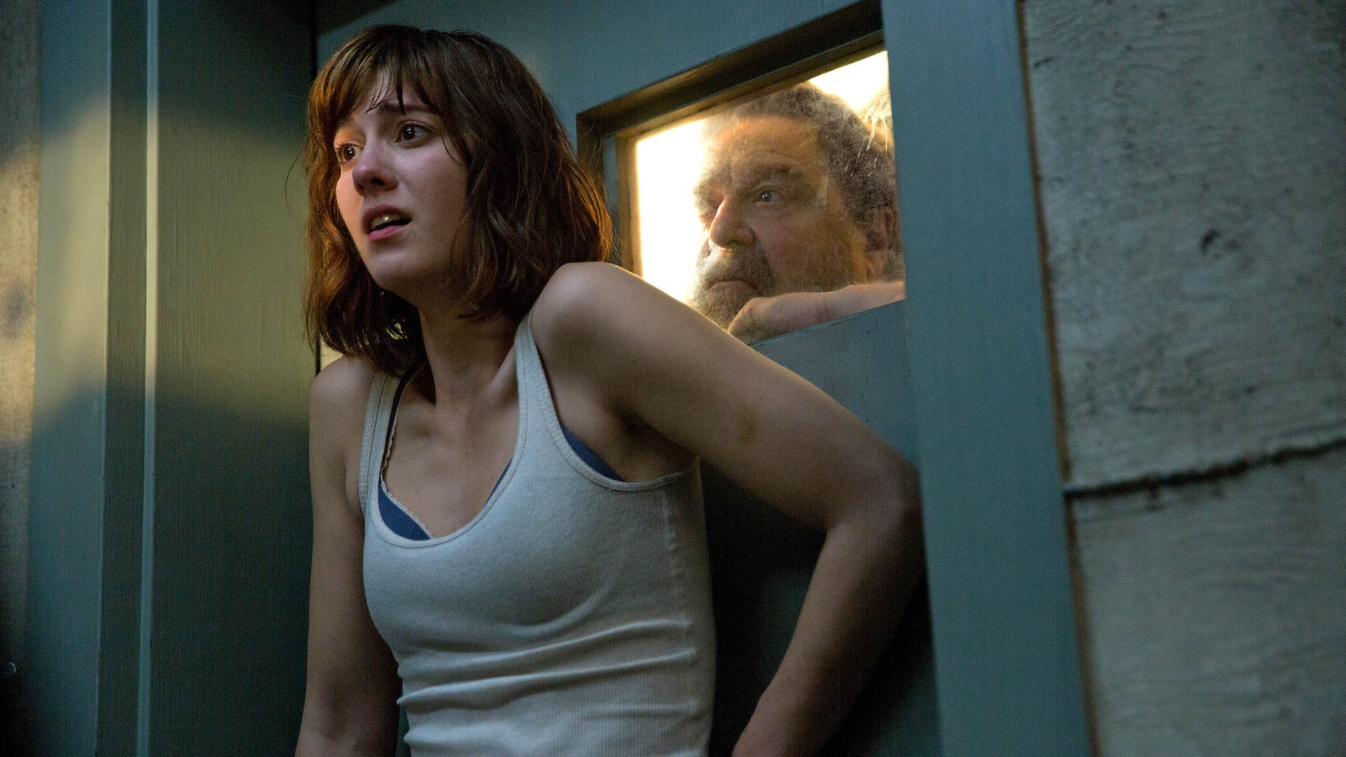 Michelle looks scared as Howard peers at her through a small window in 10 Cloverfield Lane