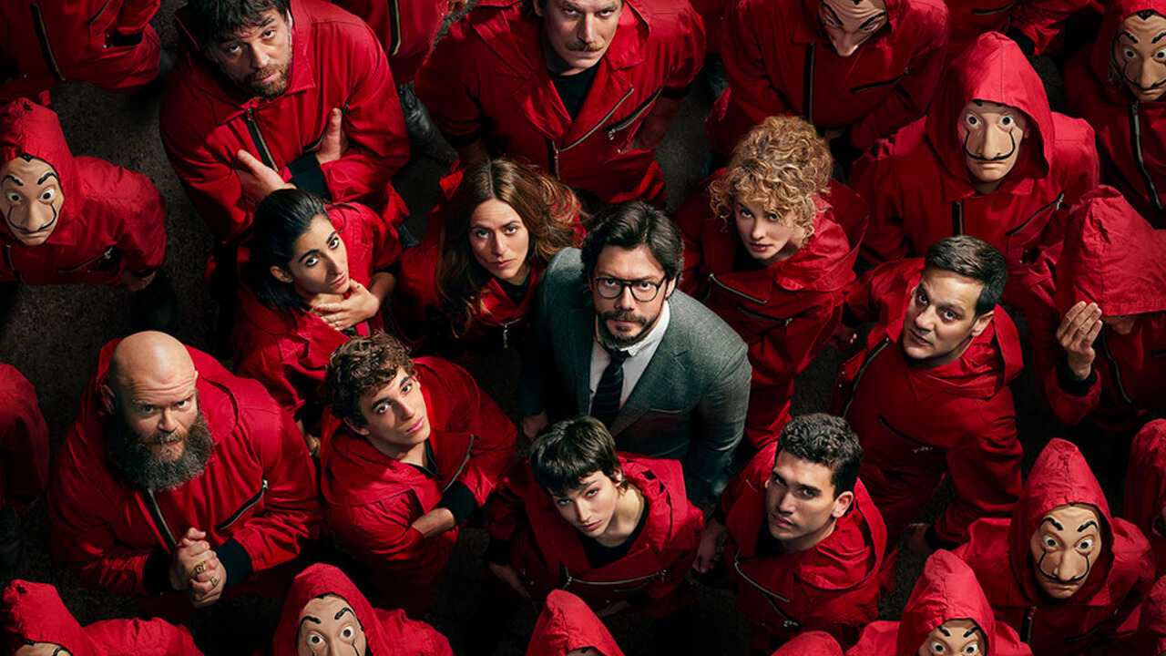 The Professor stands in the middle of the cast wearing red jumpsuits and looking up at the camera.