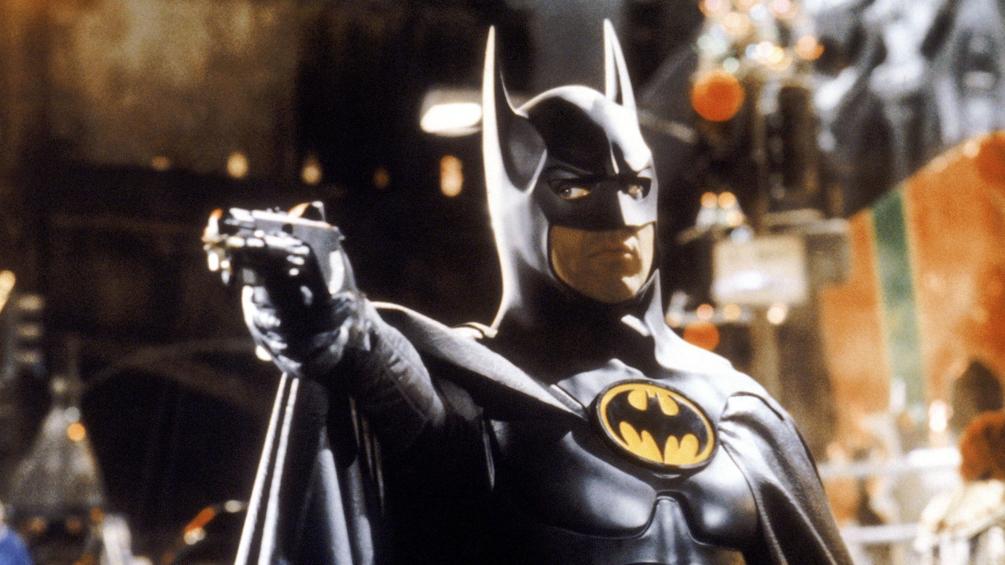 Michael Keaton as Batman in Batman Returns wearing the Batman cape and hood and pointing a gun just off camera.