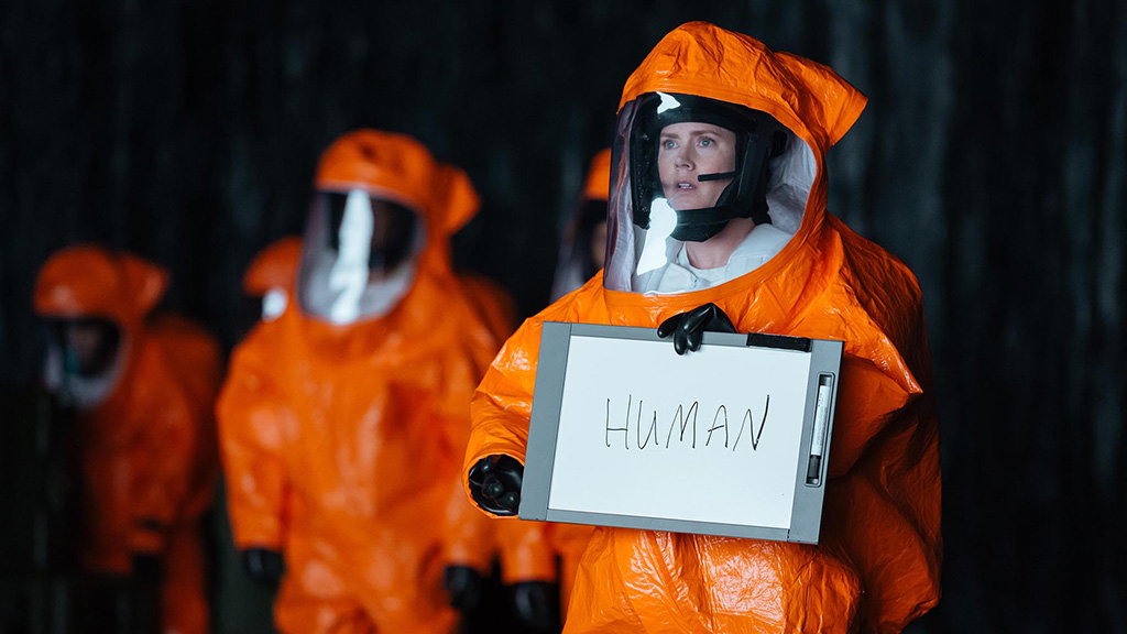 Amy Adams as Louise Banks wearing an orange hazmat suit and holding a sign with 'human' written on it in Arrival.